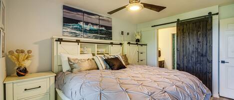 A bedroom with a bed, nightstand, dresser, ceiling fan, and artwork on the wall.