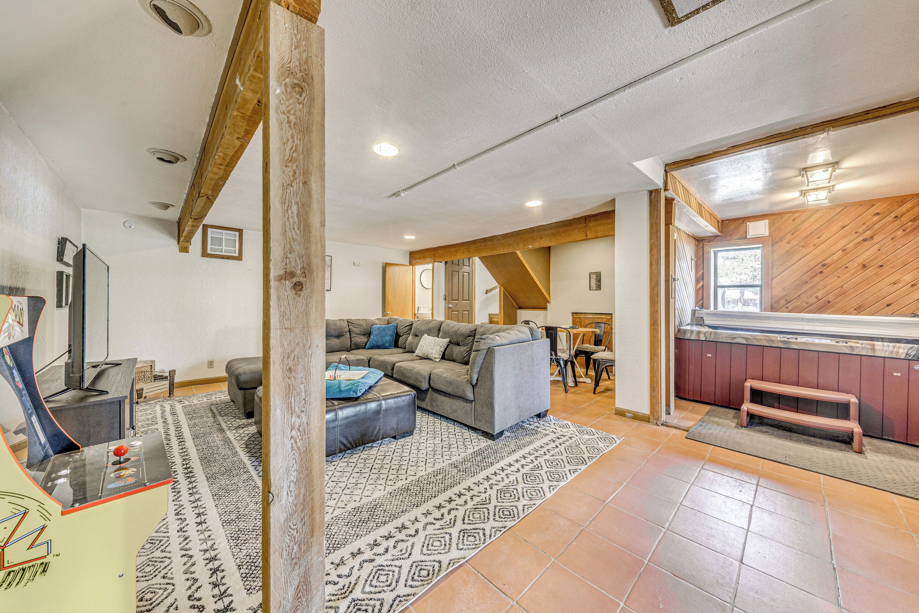 A spacious living area with a large sofa, a coffee table, and a game console. There is a wooden bench and a small table with chairs. The room has a tiled floor and a patterned rug.