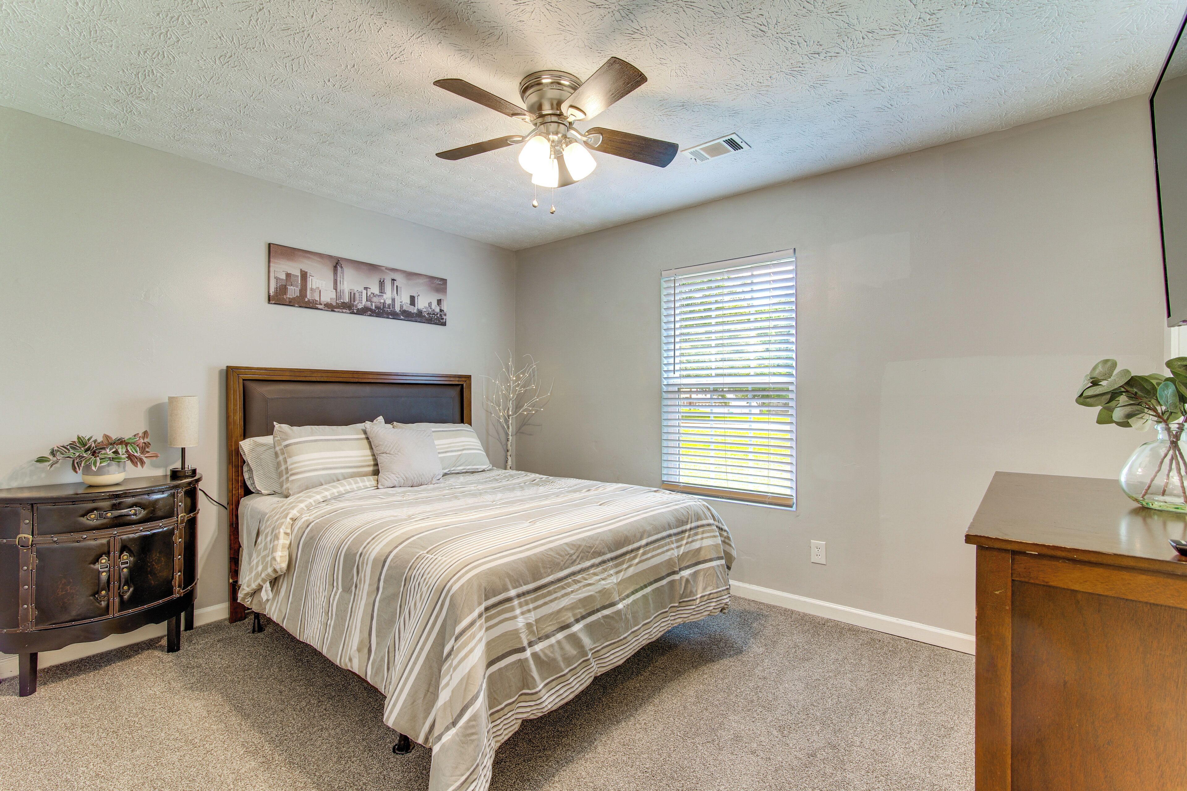 A bedroom with a bed, a ceiling fan, a nightstand, a dresser, a window with blinds, and a cityscape painting on the wall.
