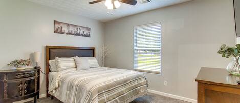 Un dormitorio con cama, ventilador de techo, mesita de noche, cómoda, ventana con persianas y una pintura de paisaje urbano en la pared.