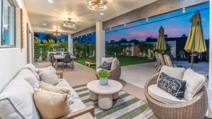 An outdoor patio with a sofa, round table, and wicker chairs. There is a dining table and chairs in the background, and a view of a garden and sunset. - NEW The Hensley on Hubbell Designer Oasis w Pool (Scottsdale)