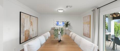 A dining area with a long wooden table, white upholstered chairs, and two large abstract paintings on the walls.
