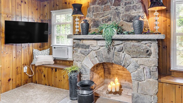 A cozy room with a stone fireplace, a mounted TV, and a comfortable seating area with a cushioned bench and a small table. - < 1 Mi to Raystown Lake: Family-friendly Cottage! (Huntingdon)
