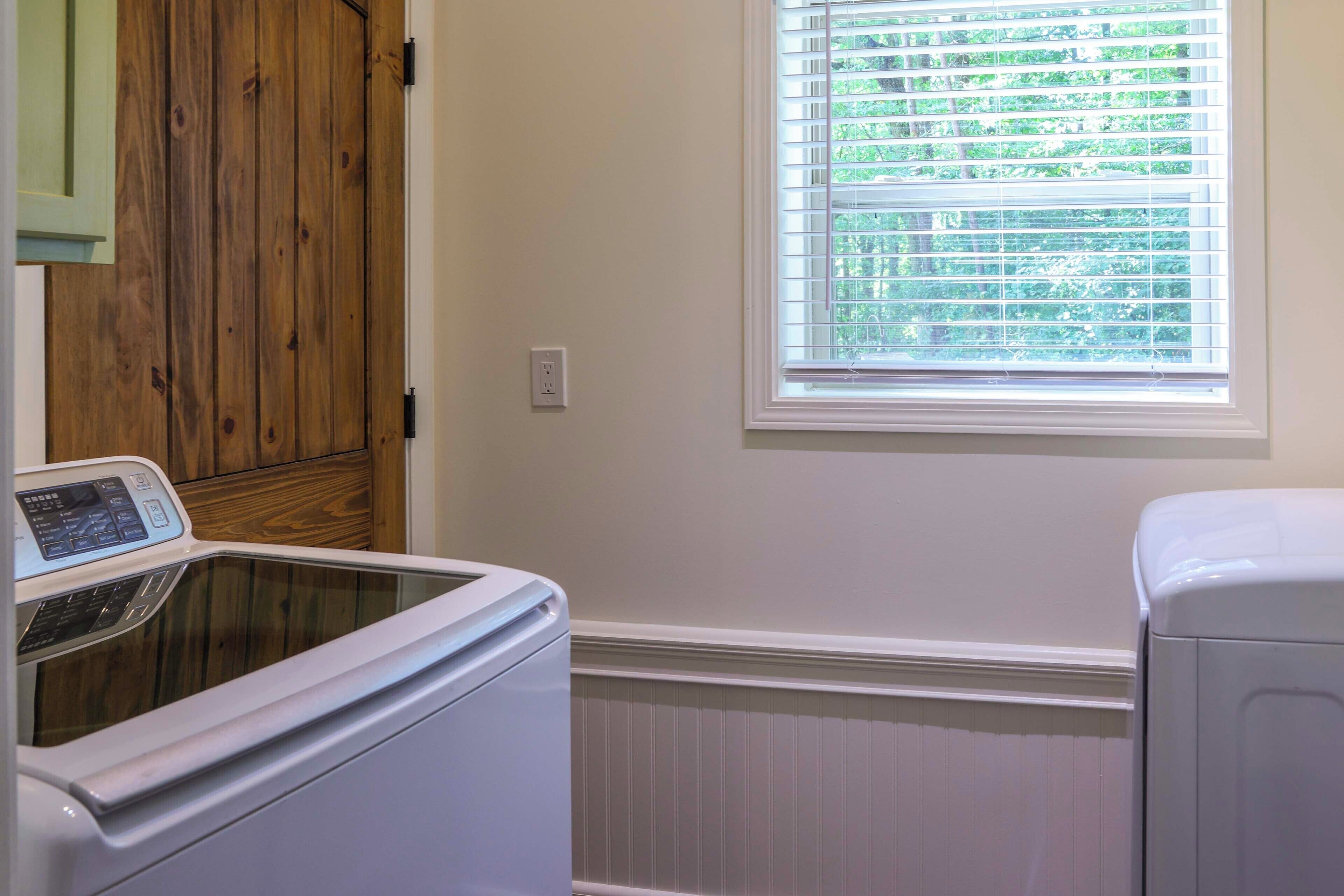A laundry room with a front-loading washer and dryer, wooden cabinets, a window with blinds, and a white wall.