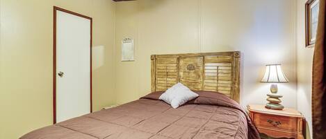 A bedroom with a wooden headboard, a single bed with a brown quilt, a nightstand with a lamp, and a door.