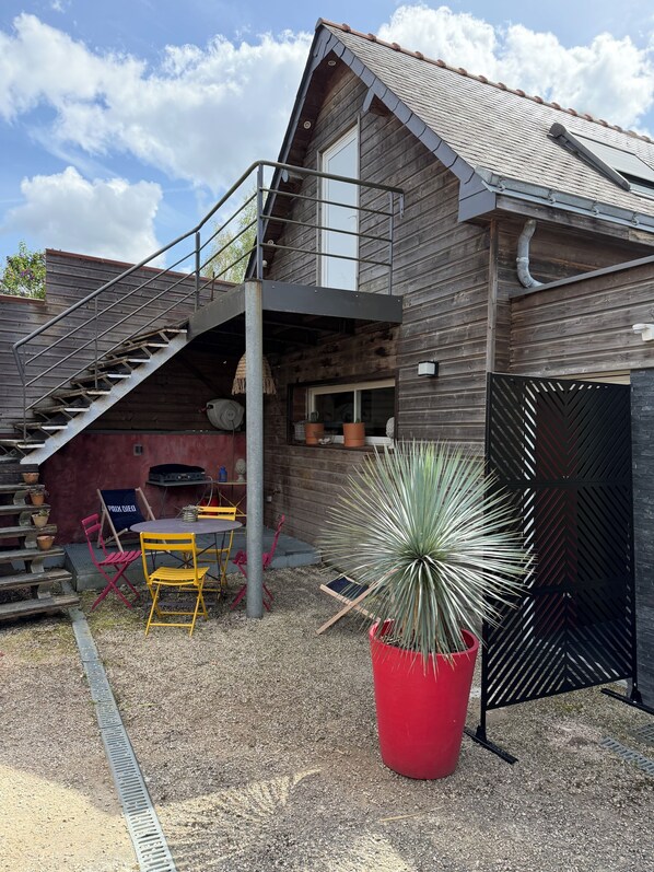 A wooden house with a staircase leading to a balcony, a patio with chairs and a table, and a large potted plant.