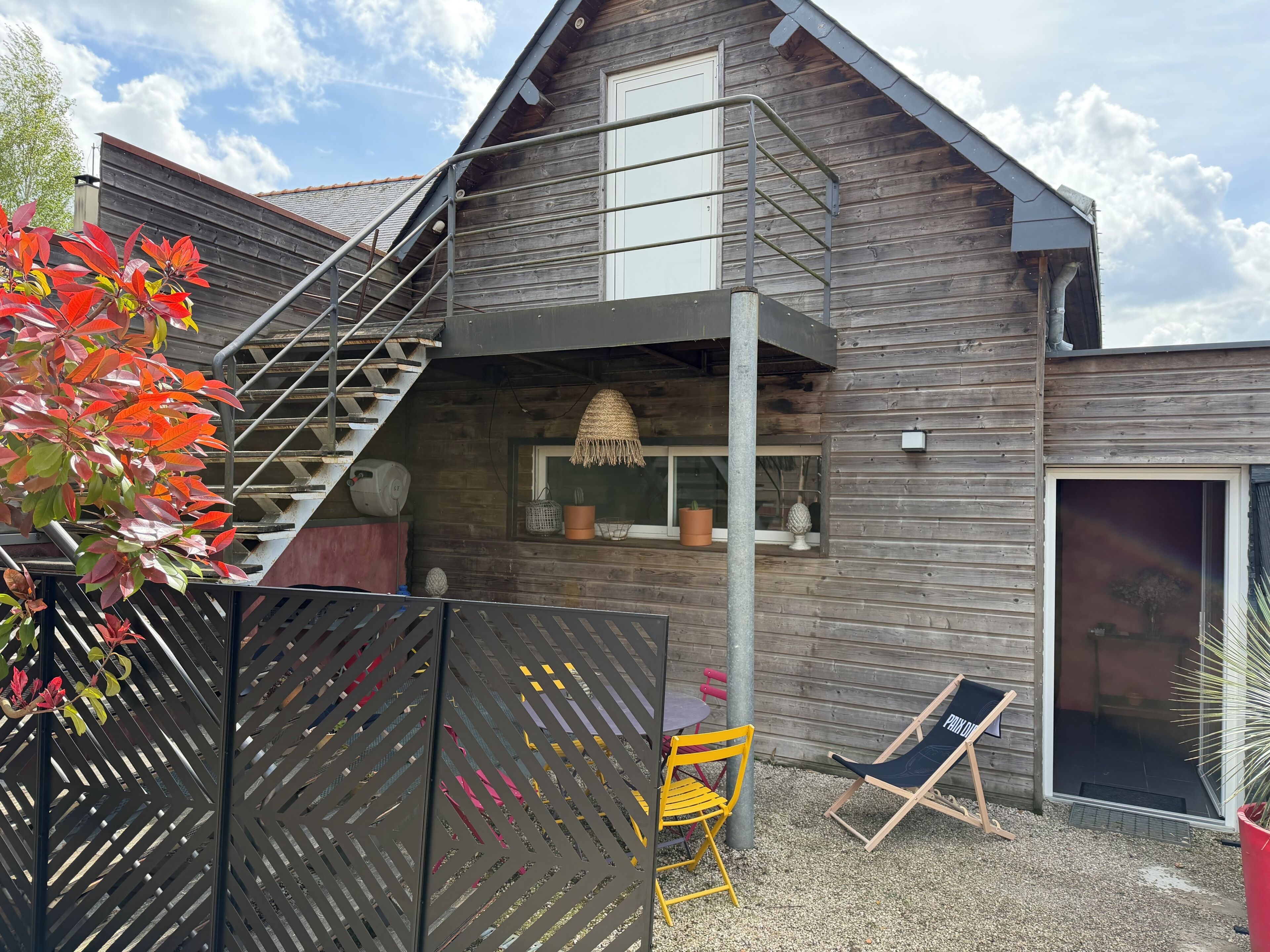 A wooden house with a balcony, a staircase, a window with a view, and outdoor seating.