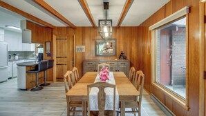 Un coin repas avec une table en bois, des chaises et un lustre. À gauche, on trouve une cuisine avec des meubles blancs et un réfrigérateur.