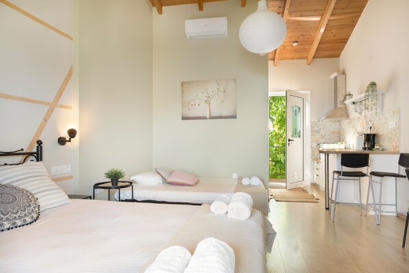 A bedroom with two beds, a small table with a plant, a painting on the wall, and a kitchenette area with bar stools.