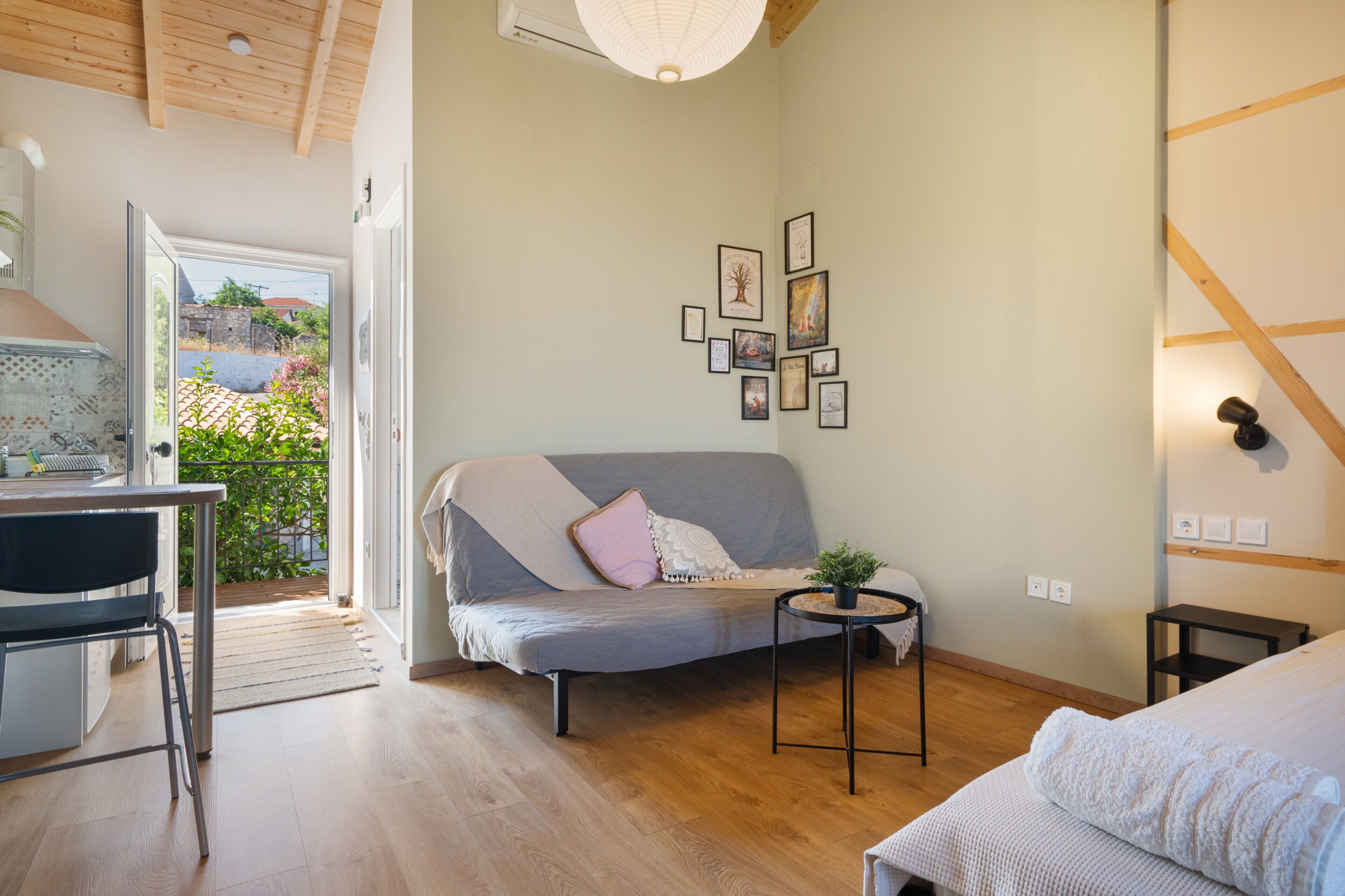 Et moderne værelse med træloft, en grå sofa med en lyserød pude, et lille rundt bord med en plante og en seng med et hvidt tæppe.