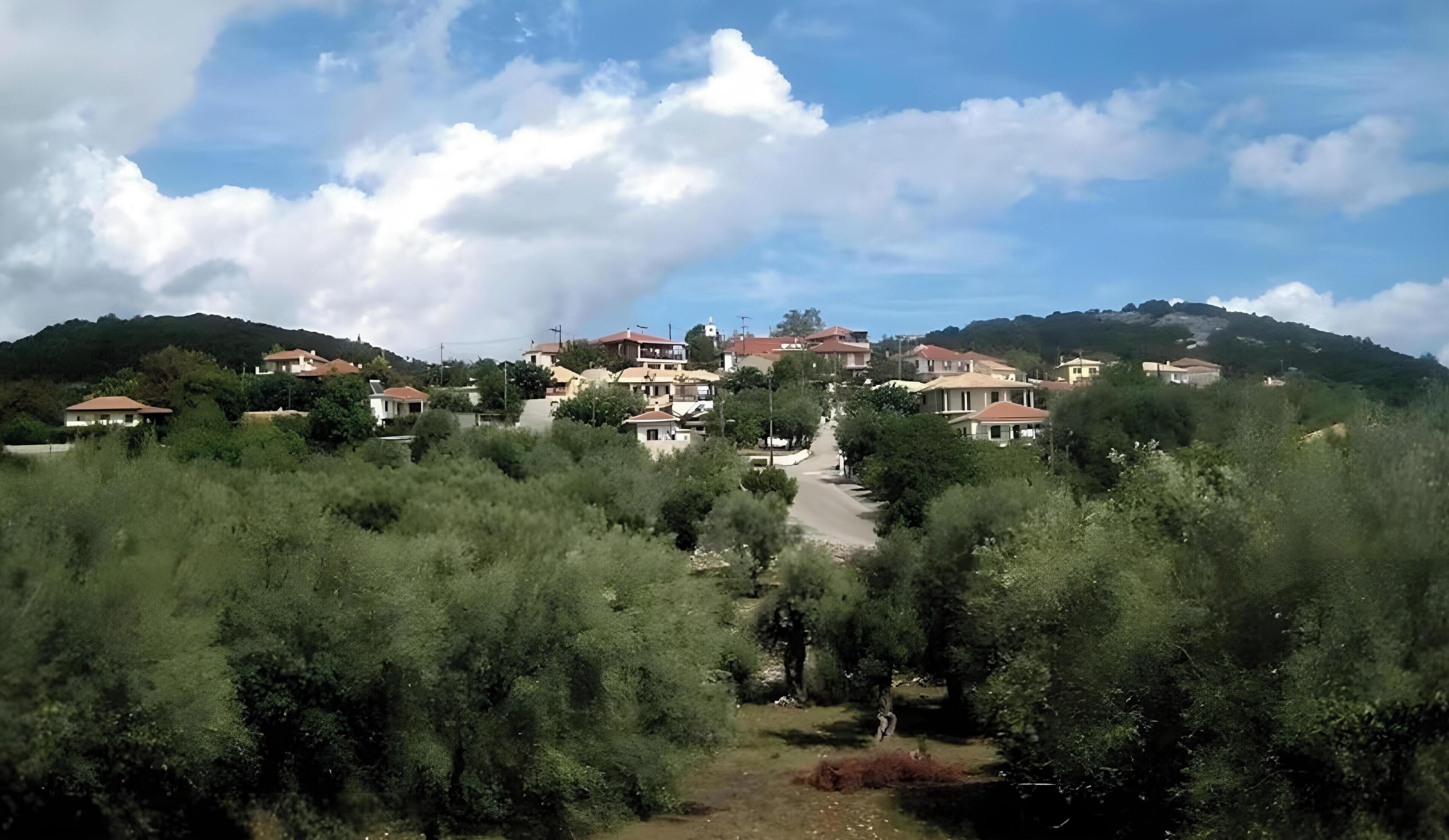 A residential area with houses on a hillside, surrounded by trees, and a clear sky.