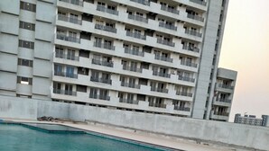 Piscina com escada de metal, cercada por um deck de concreto e um edifício alto ao fundo.