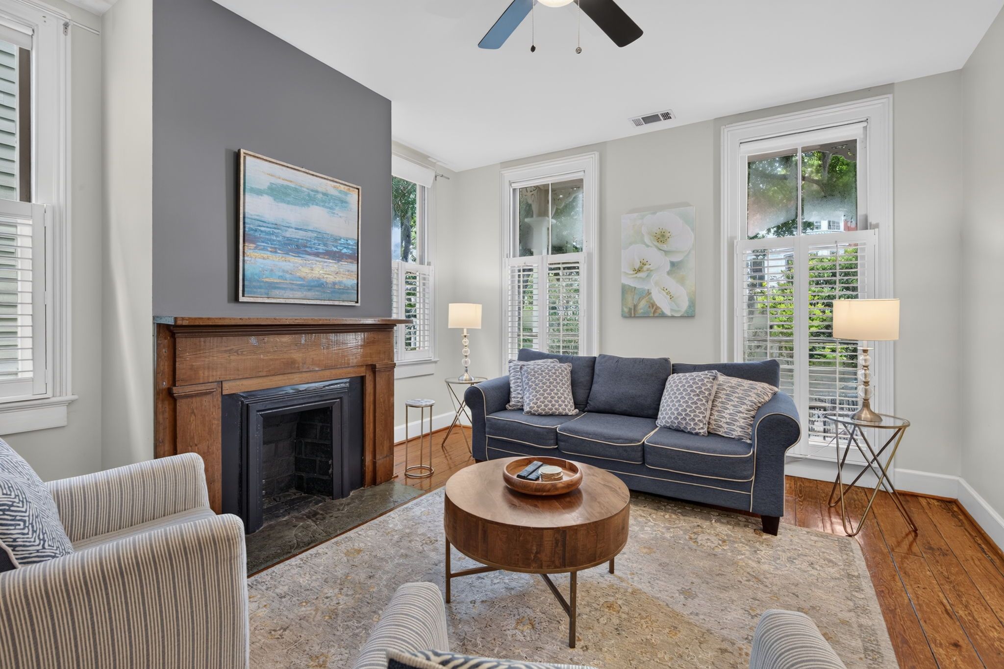 A living room with a fireplace, a sofa, a coffee table, and a view of greenery through the windows.