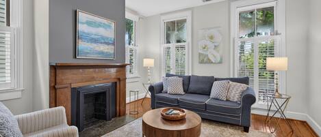 A living room with a fireplace, a sofa, a coffee table, and a view of greenery through the windows.