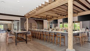 Zona de bar moderna con vigas de madera, una barra larga con taburetes y un gran ventanal circular.