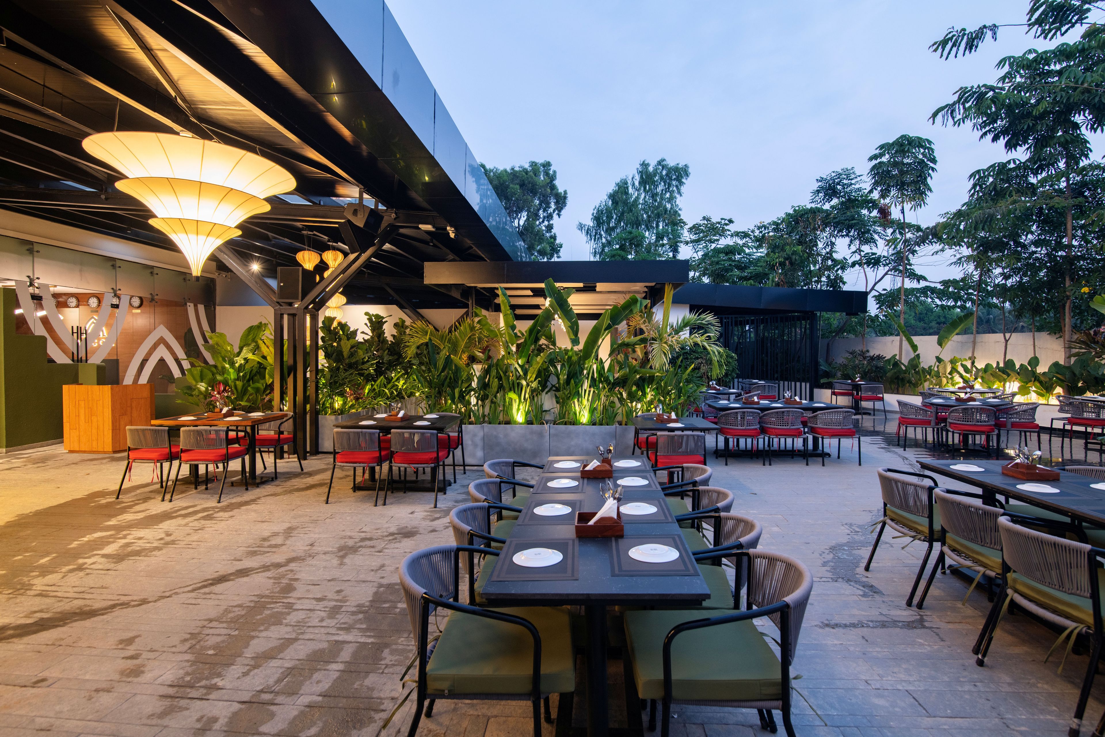 An outdoor dining area with tables and chairs, surrounded by greenery and a modern building structure.