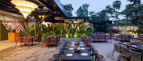 An outdoor dining area with tables and chairs, surrounded by greenery and a modern building structure.