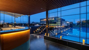 A modern indoor pool area with a bar and seating area, large windows, and a view of the cityscape.