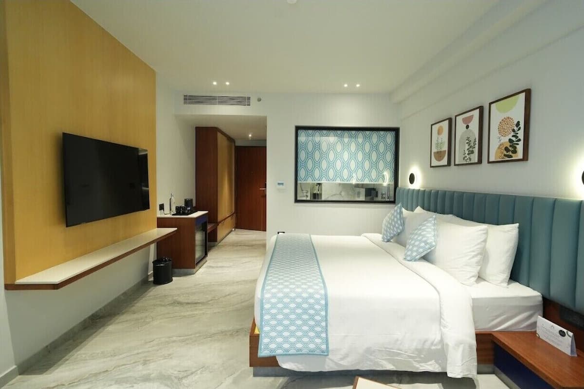 A hotel room with a bed, a TV, a desk, and a bathroom visible through a glass door.