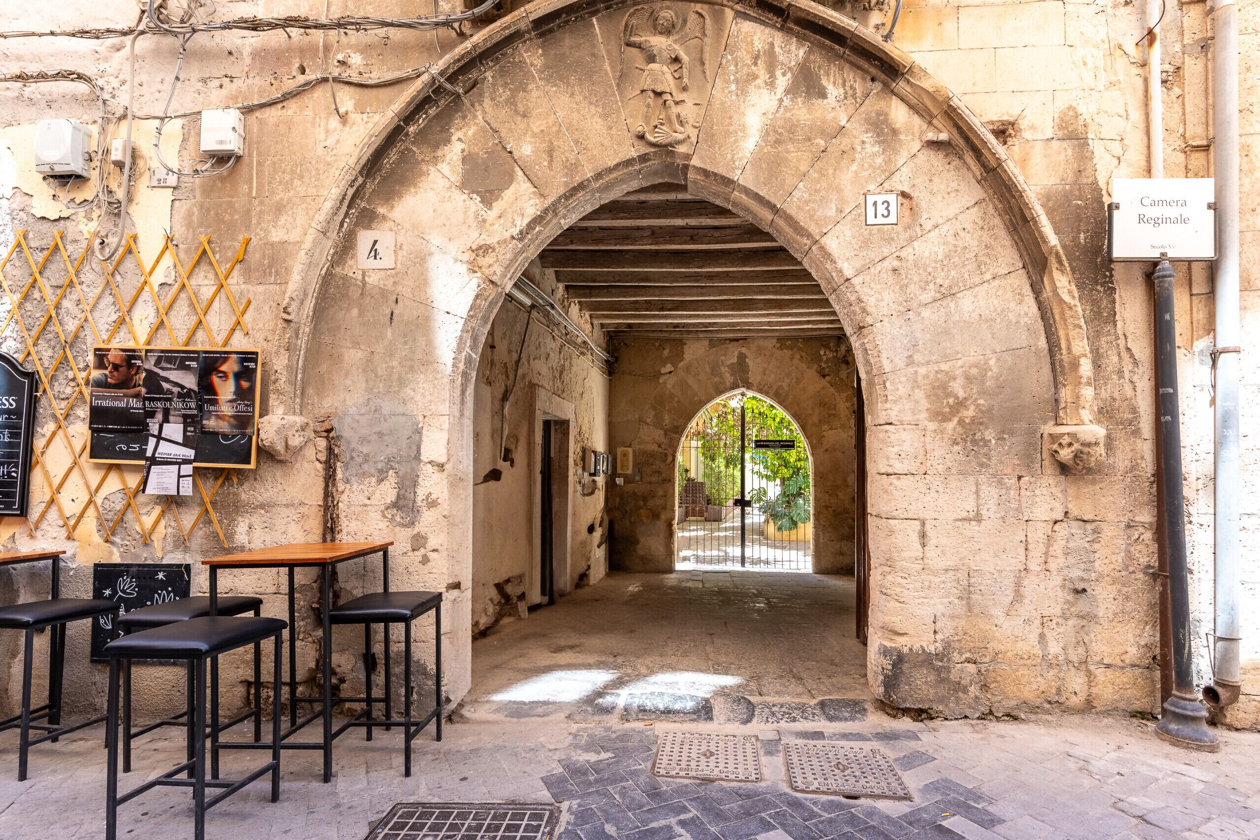 Un arco de piedra con un cartel con el número 13, que da a un patio con una puerta. Hay mesas y sillas al aire libre a la izquierda.