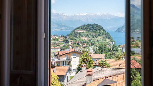 A view from a window overlooking a hillside village with buildings, a lake, and mountains in the background.