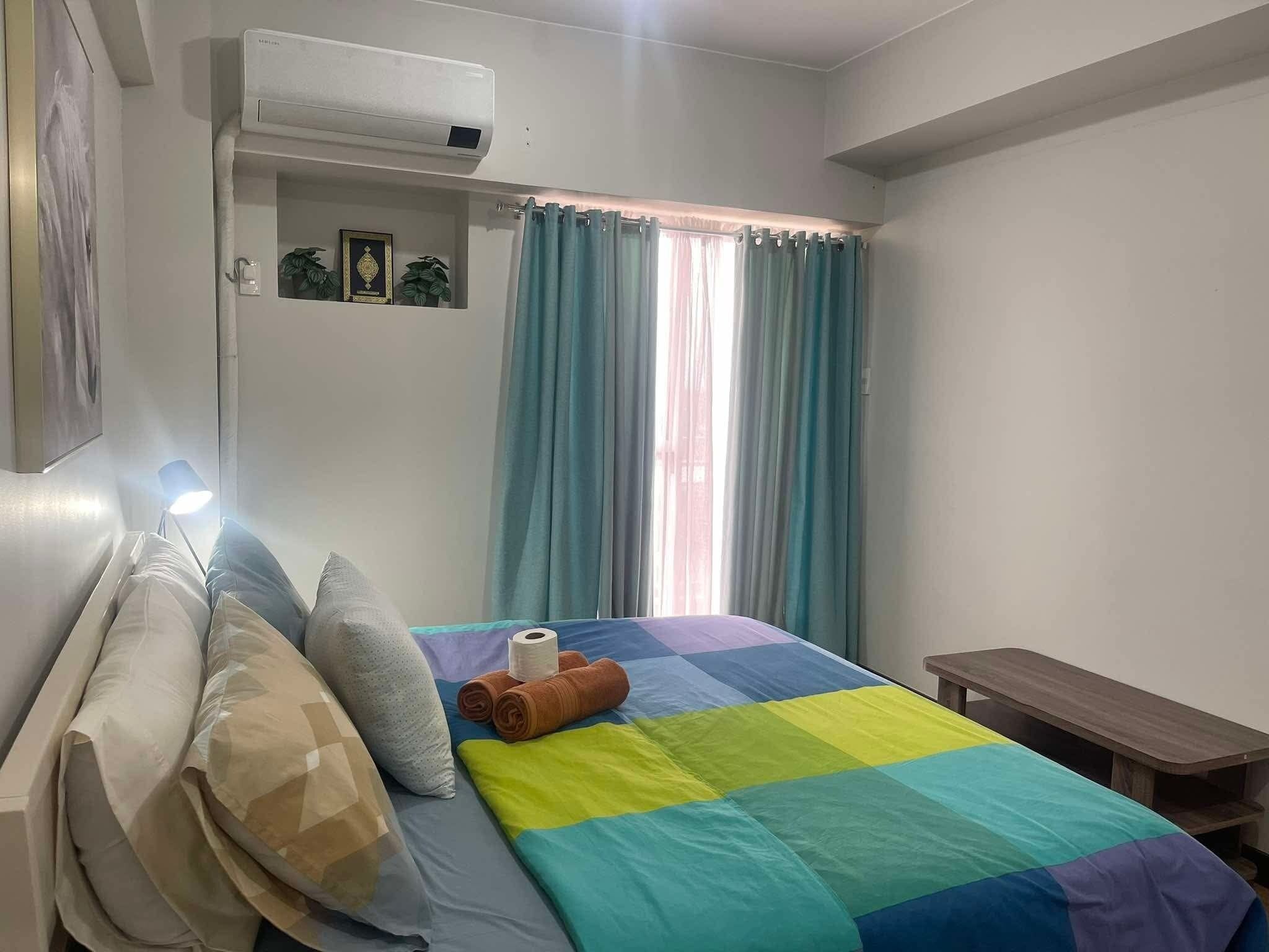 Una camera da letto con un letto, cuscini, un comodino, una lampada, una finestra con tende e un condizionatore d'aria.