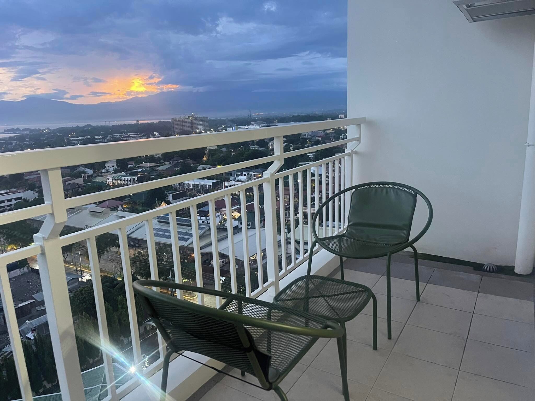A balcony with two green chairs, a white railing, and a view of a cityscape during sunset.