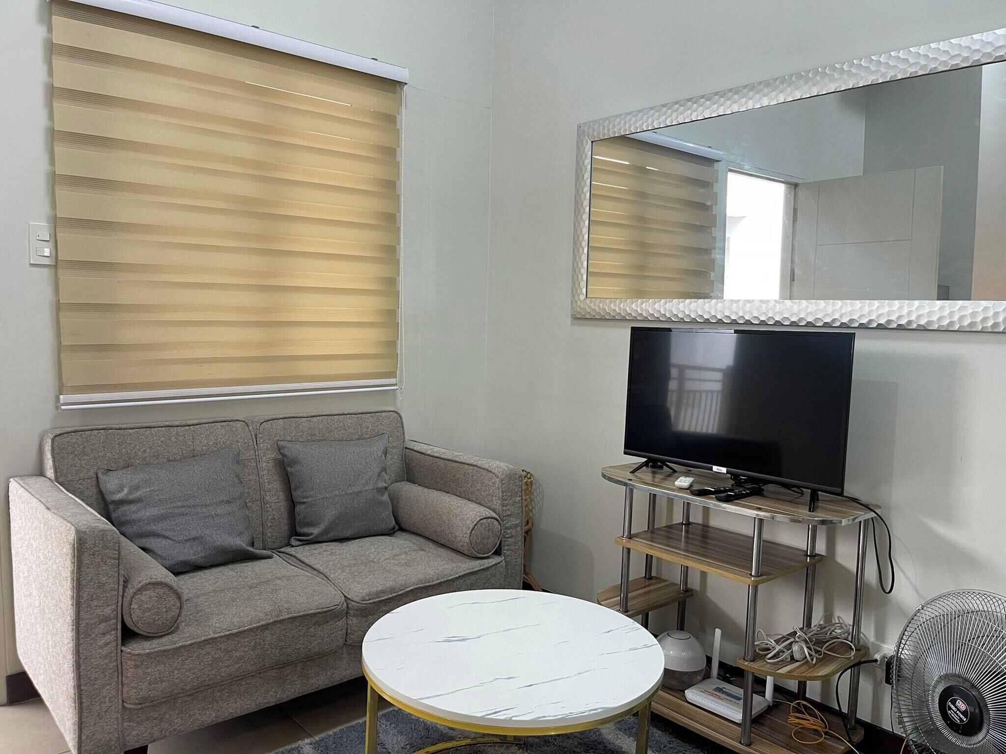 A living room with a grey sofa, a white round coffee table, a TV on a metal stand, and a large mirror above the TV.