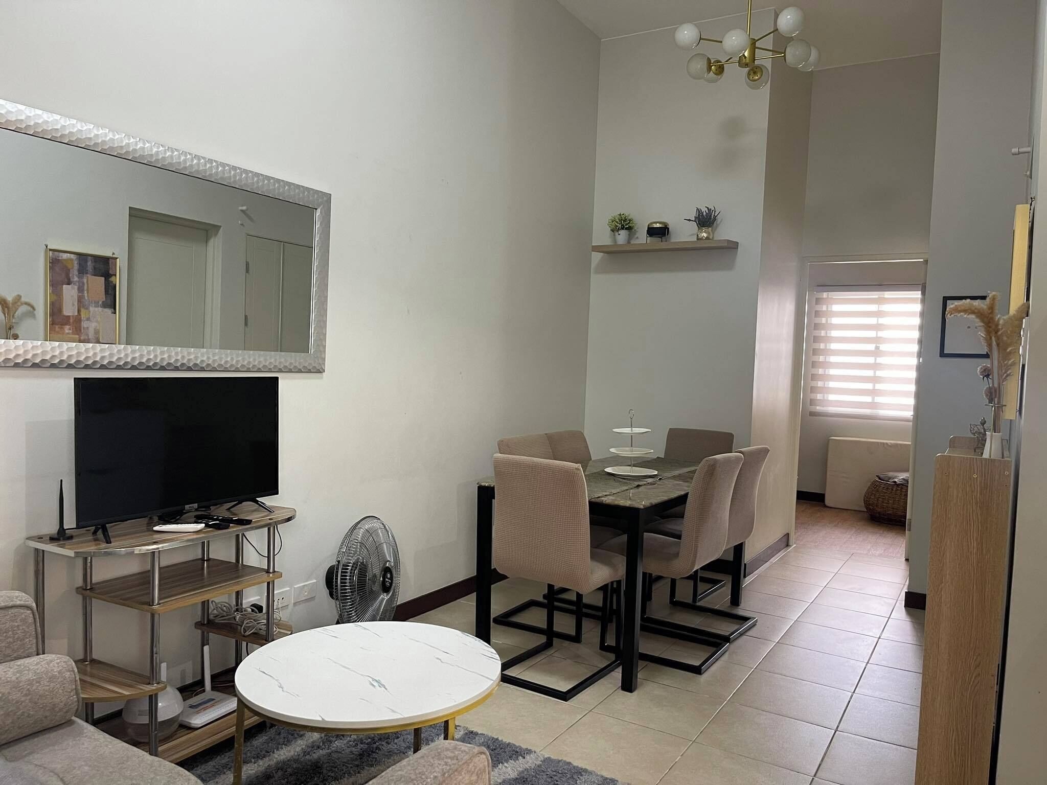 A modern living room with a flat-screen TV, a dining table with chairs, a standing fan, and a large mirror.