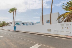 Uma estrada costeira com um edifício branco com detalhes azuis, palmeiras e céu claro.