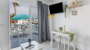A room with a balcony, a TV, a table with chairs, and a small shelf with decorative items.