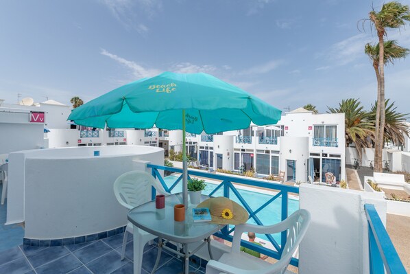 Área da piscina com um guarda-sol turquesa, cadeiras brancas e uma mesa com chapéu e livro.