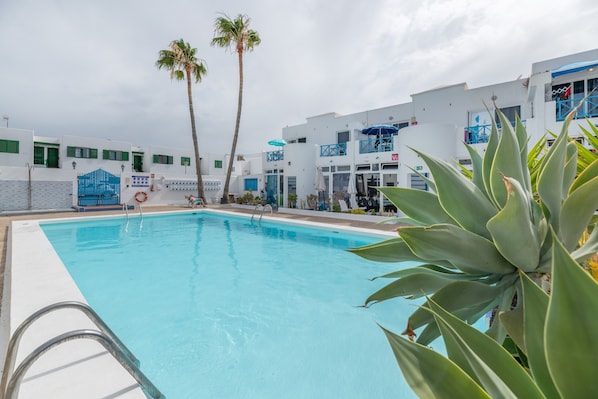 A swimming pool with a clear blue water, surrounded by white buildings with blue accents and palm trees. - Sueño Del Mar Vacation Apartment with Shared Pool, Wi-Fi & Air Conditioning (Puerto del Carmen)