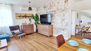 A modern living room with a wooden floor, a television mounted on a textured wall, a desk with a chair, and a dining area with blue plates.