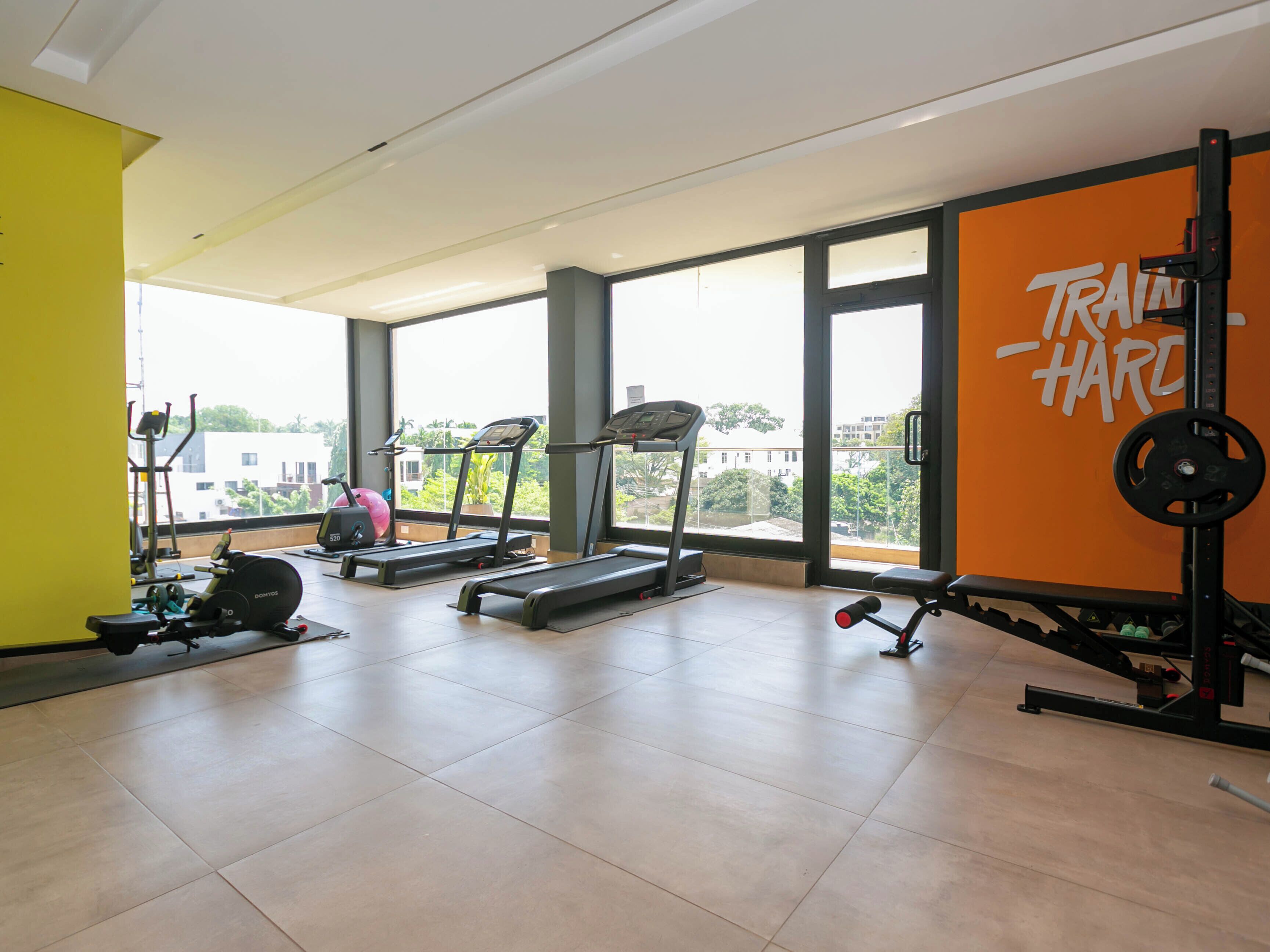 Een fitnessruimte met loopbanden, een gewichtbank en een muur waarop 'TRAIN HARD' staat.