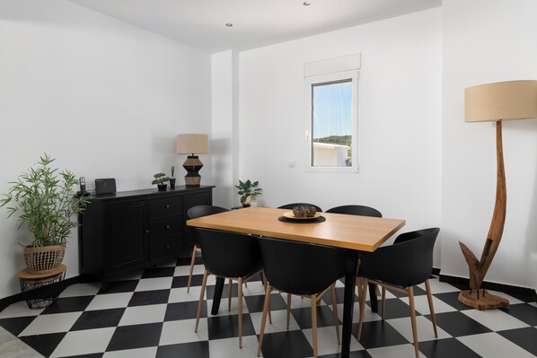 Uma área de refeições com piso xadrez, uma mesa de madeira, cadeiras pretas, um armário preto, um abajur e uma janela com vista.