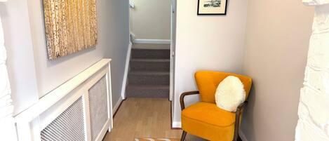 A hallway with a radiator, a patterned rug, a chair with a fluffy pillow, and a staircase leading to another room.