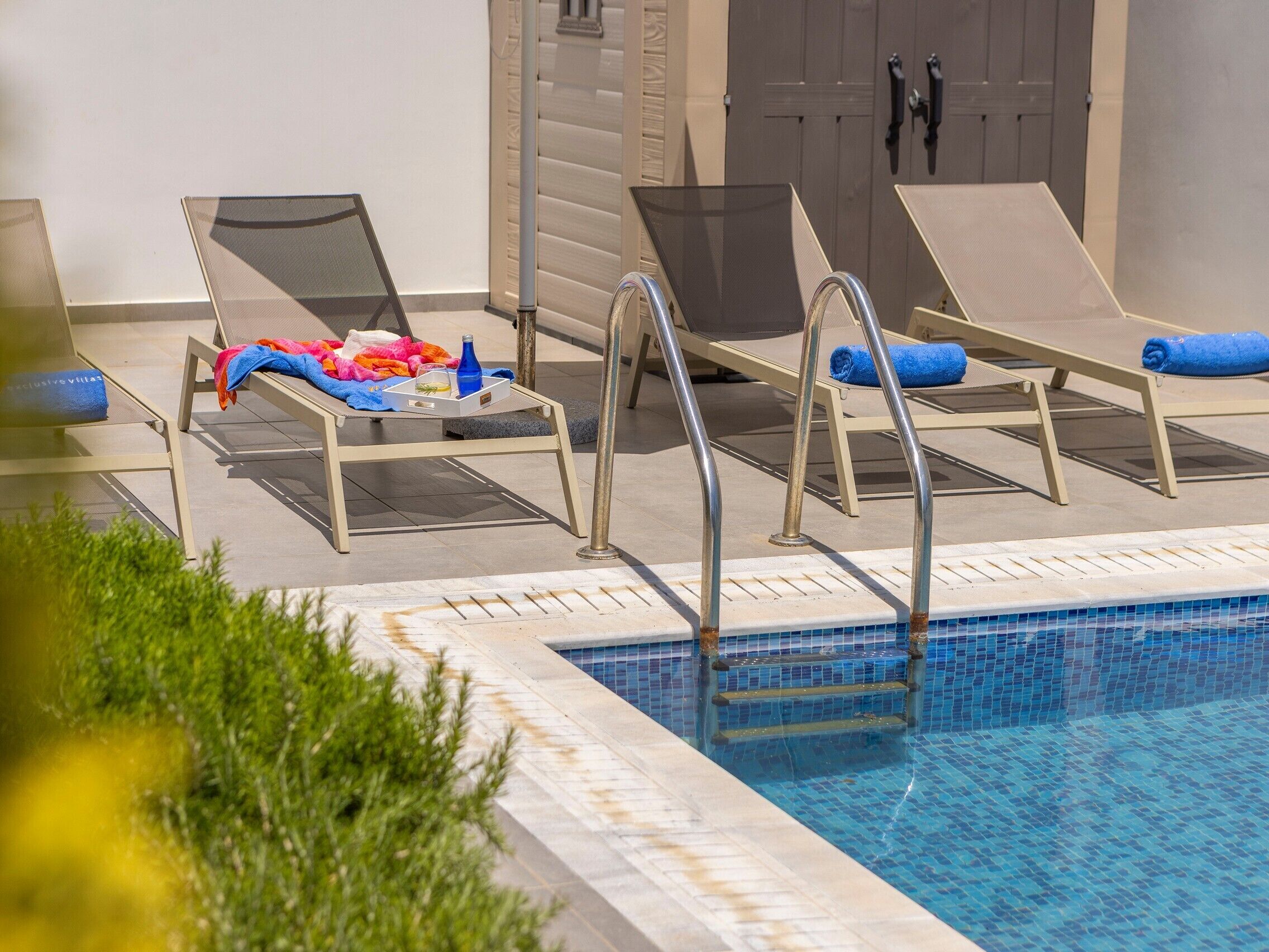 A poolside area with lounge chairs, a table with towels and a bottle, and a door in the background.