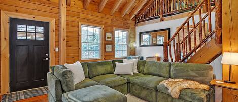 Interior acogedor de una cabaña con un sofá seccional verde, paredes de madera, una escalera y un gran ventanal.