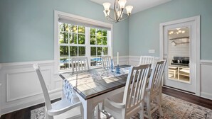 Ein Esszimmer mit großem Tisch, weißen Stühlen, einer Chandelier und Blick auf einen Hinterhof mit Pool.