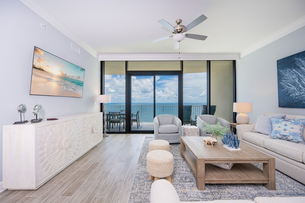 A modern living room with a balcony, a ceiling fan, a white console table, and a large painting of a beach scene.