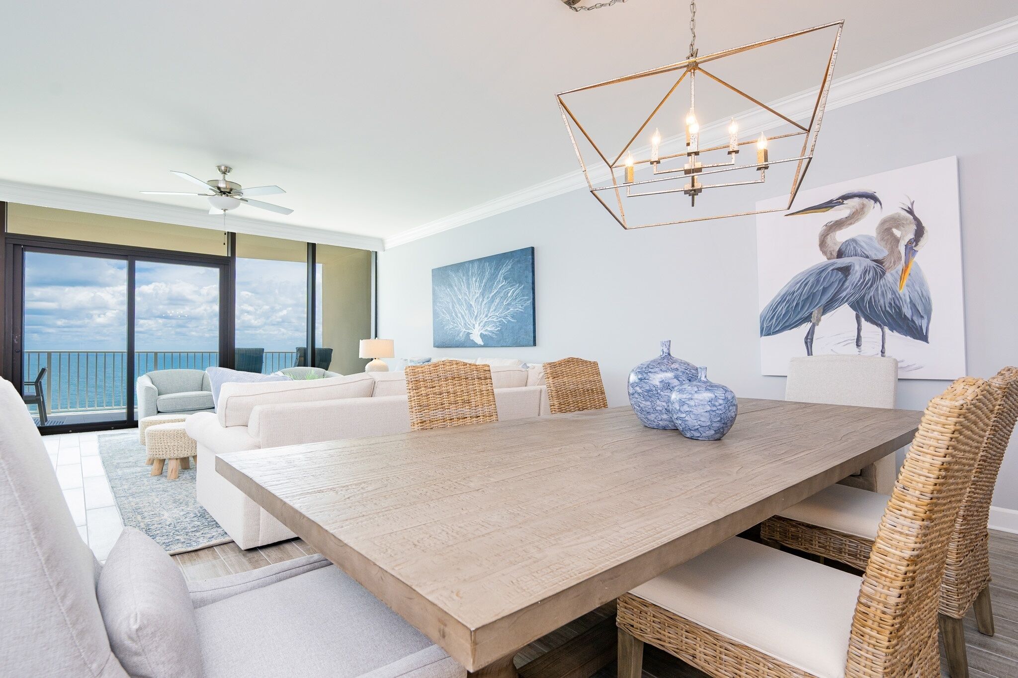 A spacious living room with a large wooden dining table, white sofas, and wicker chairs. There is a balcony with a view of the ocean, a ceiling fan, and a modern chandelier.