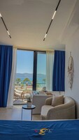 Une chambre d’hôtel moderne avec une grande fenêtre, un lit, une chaise, une petite table et un attrape-rêves accroché au mur.