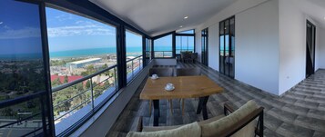 Un balcón con mesa y sillas de madera, ventanas amplias con vistas al mar y al paisaje urbano.