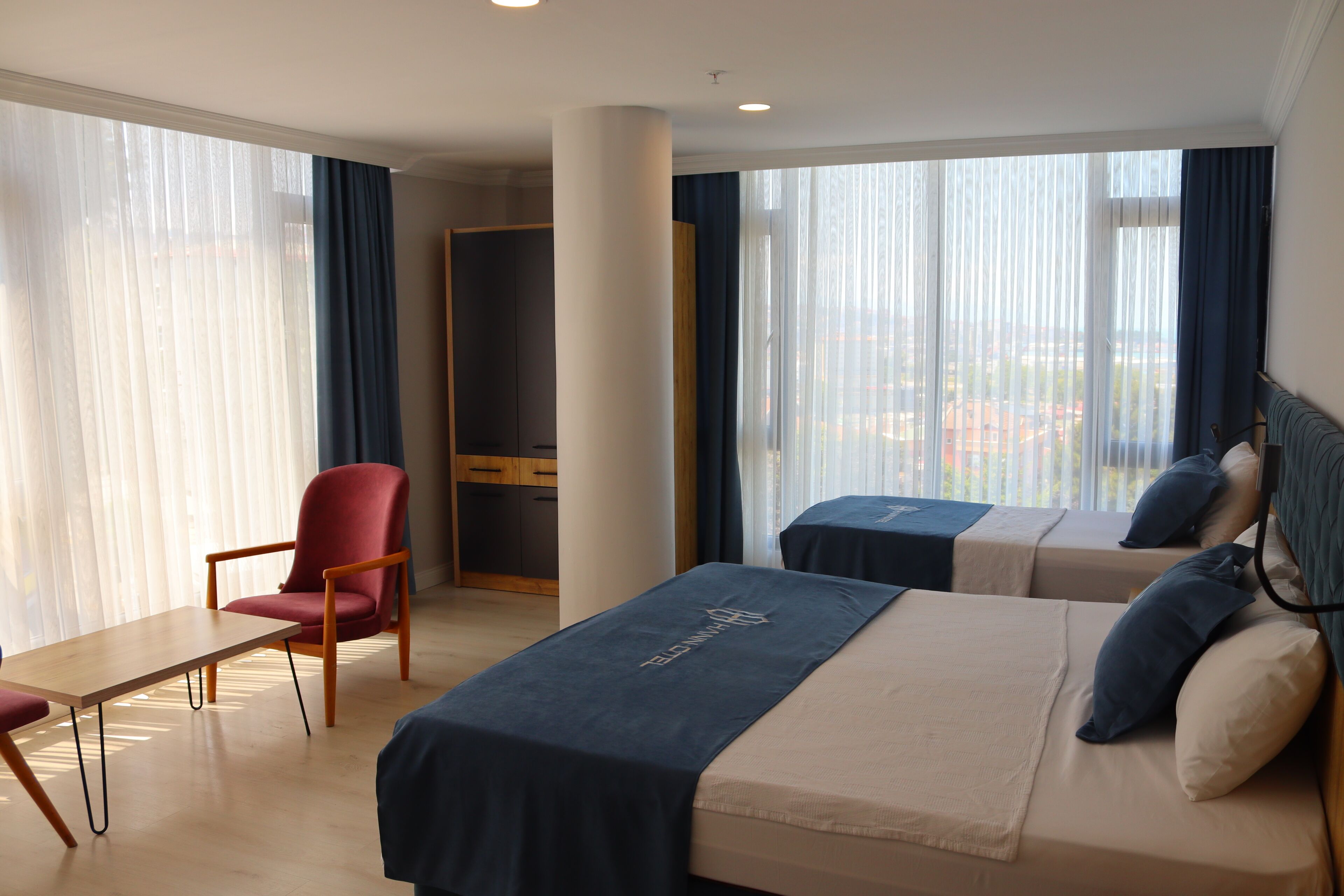 Ett hotellrum med två sängar, ett skrivbord, en stol och stora fönster med gardiner.