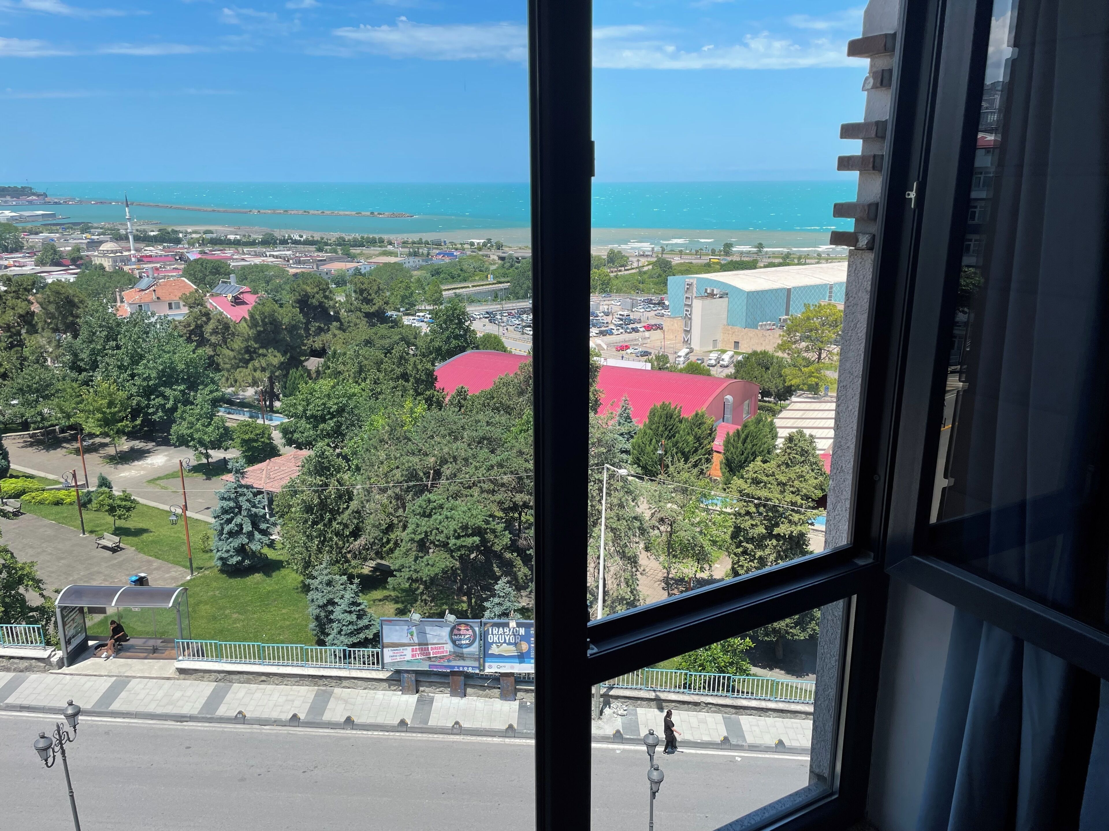Une vue depuis une fenêtre donnant sur une ville côtière, avec un mélange de bâtiments résidentiels et commerciaux, un ciel bleu dégagé et une vue lointaine sur la mer.
