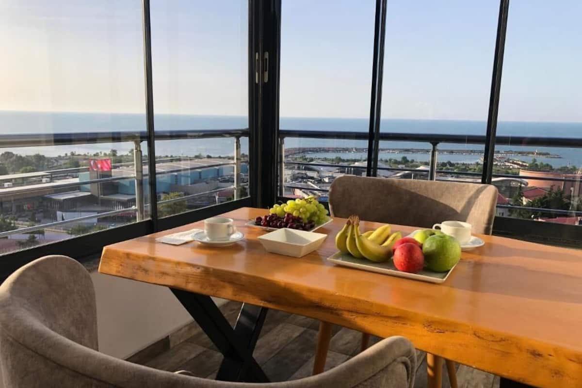 A dining table with fruits and cups, a view of the sea and cityscape through large windows.