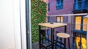 Un petit balcon avec une table en bois et deux tabourets, un mur recouvert de verdure et une vue sur un immeuble en briques.