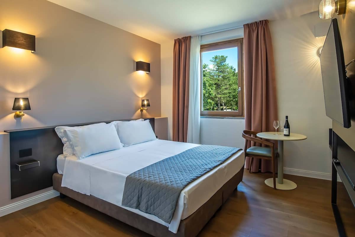 A hotel room with a bed, bedside lamps, a small table with a bottle and glass, a chair, and a TV.