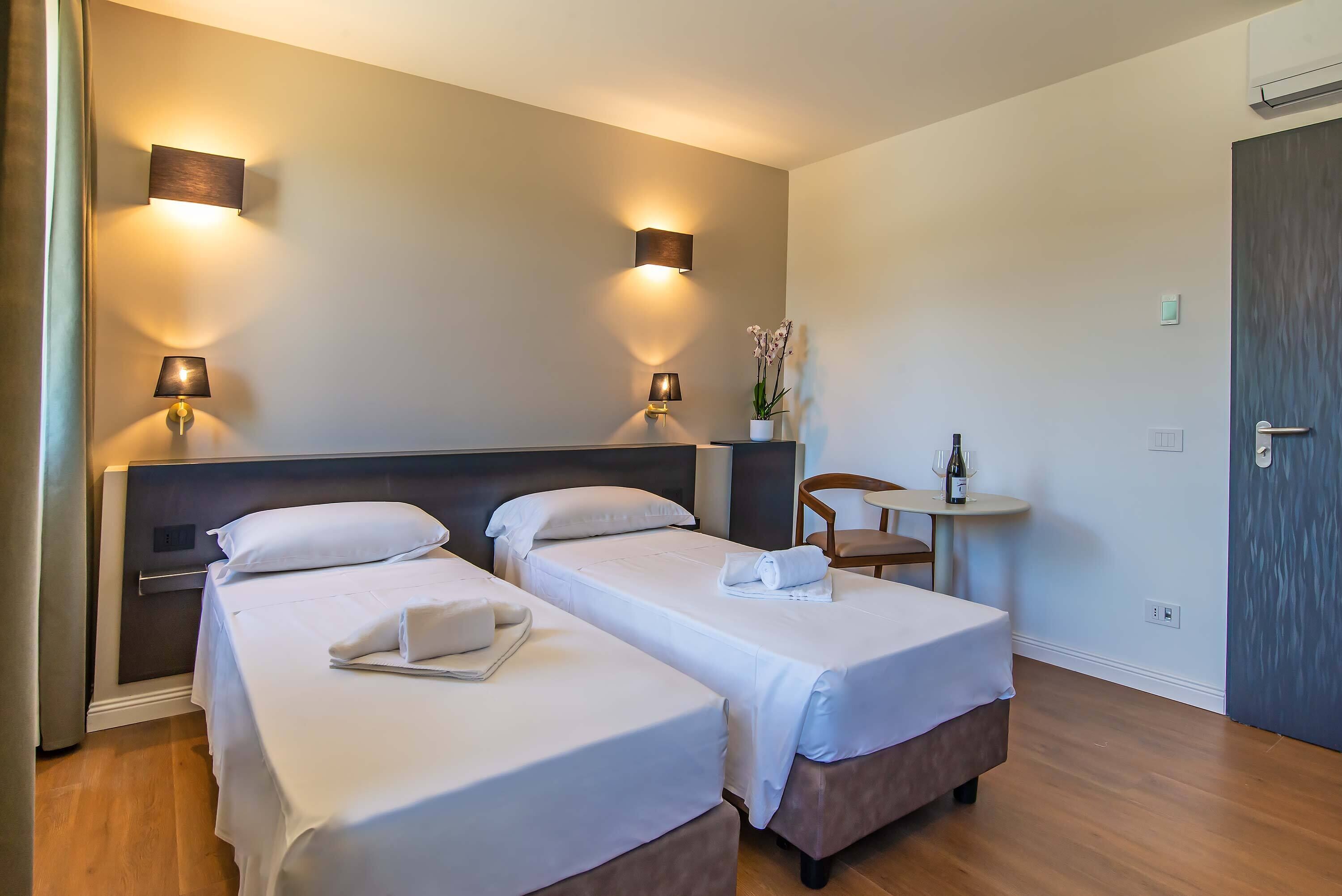 A hotel room with two single beds, bedside lamps, a small table with a chair, and a potted plant.